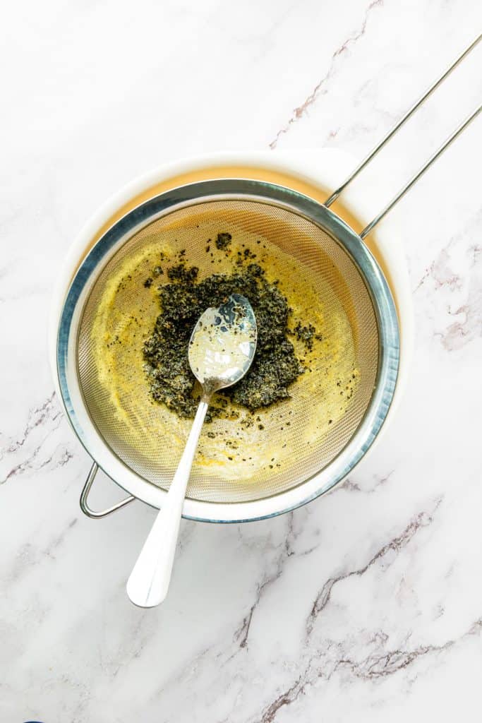 Metal sieve with green herbs and a spoon over a white bowl on a marble counter, viewed from above—perfect for preparing fresh passion fruit juice or straining ingredients for your favorite fruit juice recipes.