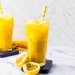 Two tall glasses of yellow passion fruit smoothie, made with refreshing passion fruit juice, sit on the counter with striped straws, ice, and passion fruit halves nearby.