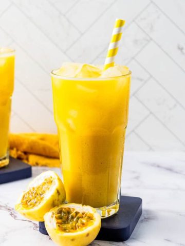 Two tall glasses of yellow passion fruit smoothie, made with refreshing passion fruit juice, sit on the counter with striped straws, ice, and passion fruit halves nearby.