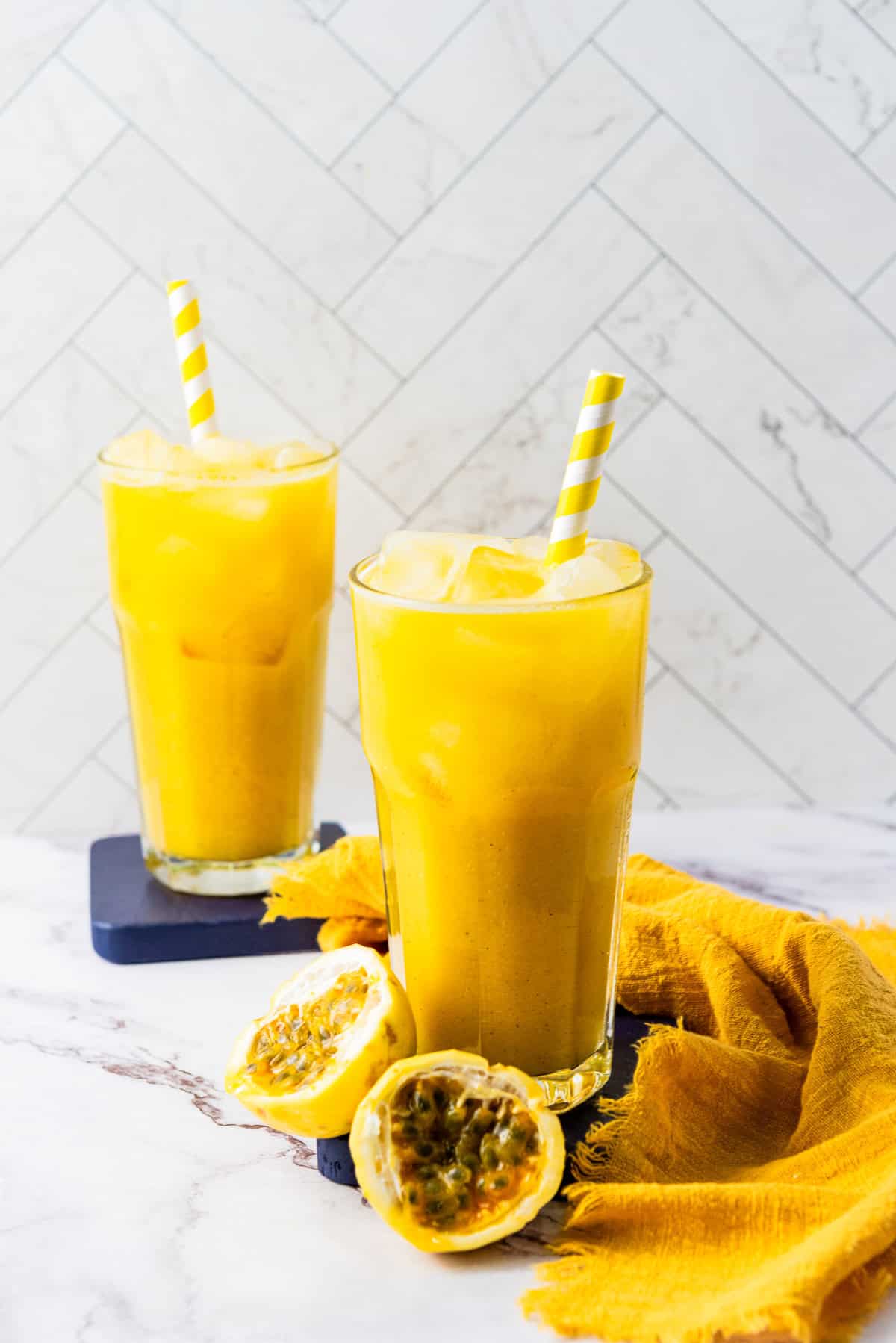 Two glasses of refreshing Passion Fruit Juice with striped straws, ice, and a halved passion fruit on a marble surface.