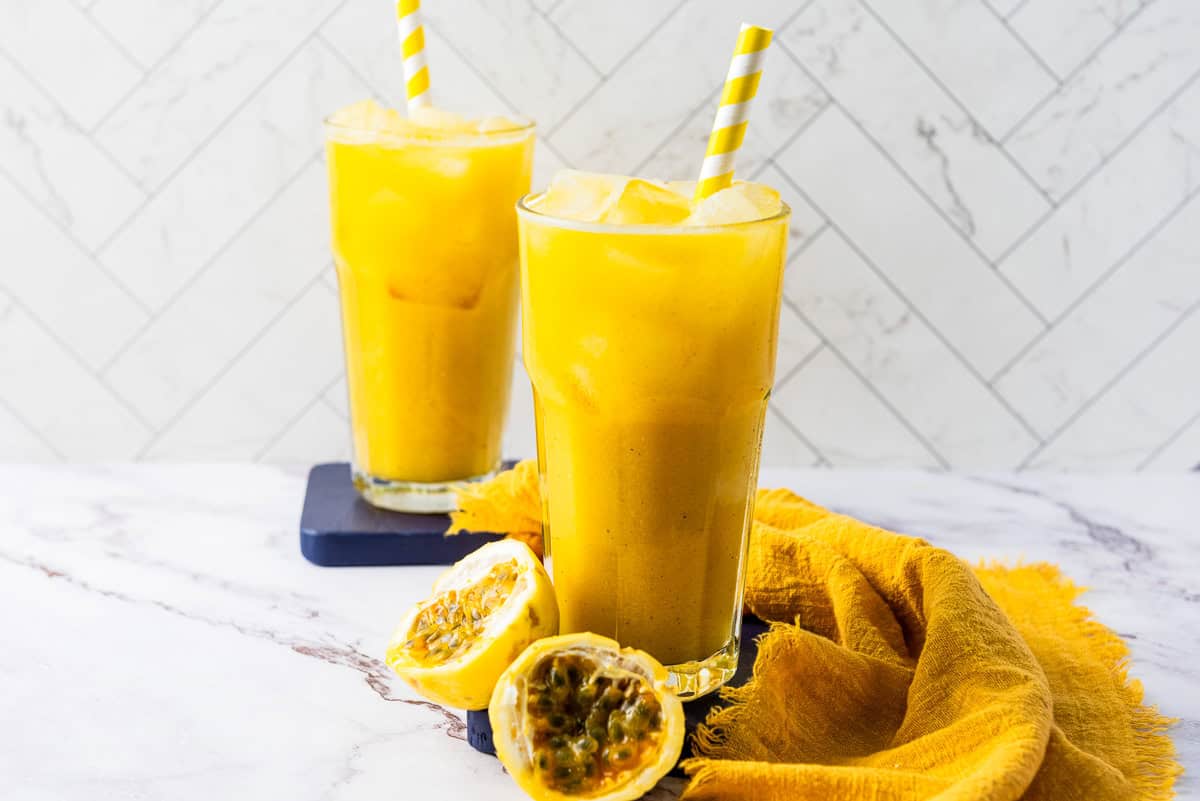 Two glasses of refreshing passion fruit juice with ice, striped straws, and half a passionfruit are arranged on a marble surface.