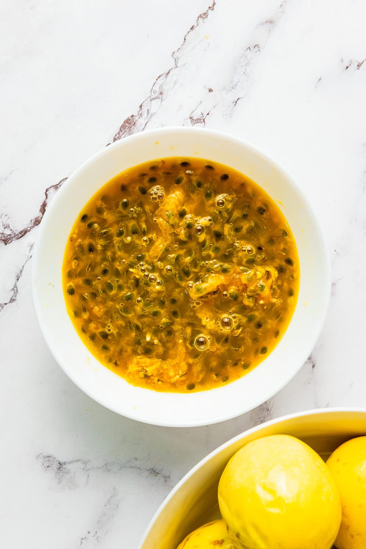 White bowl filled with yellow passion fruit pulp and seeds, perfect for making fresh passion fruit juice, sits next to lemons on a white marble surface.