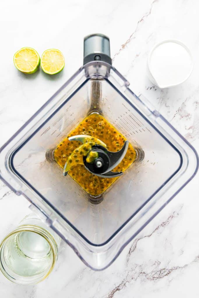 Top view of a blender with passion fruit, next to a halved lime, glass, and cup of sugar on a marble counter—perfect ingredients for making refreshing Passion Fruit Juice.