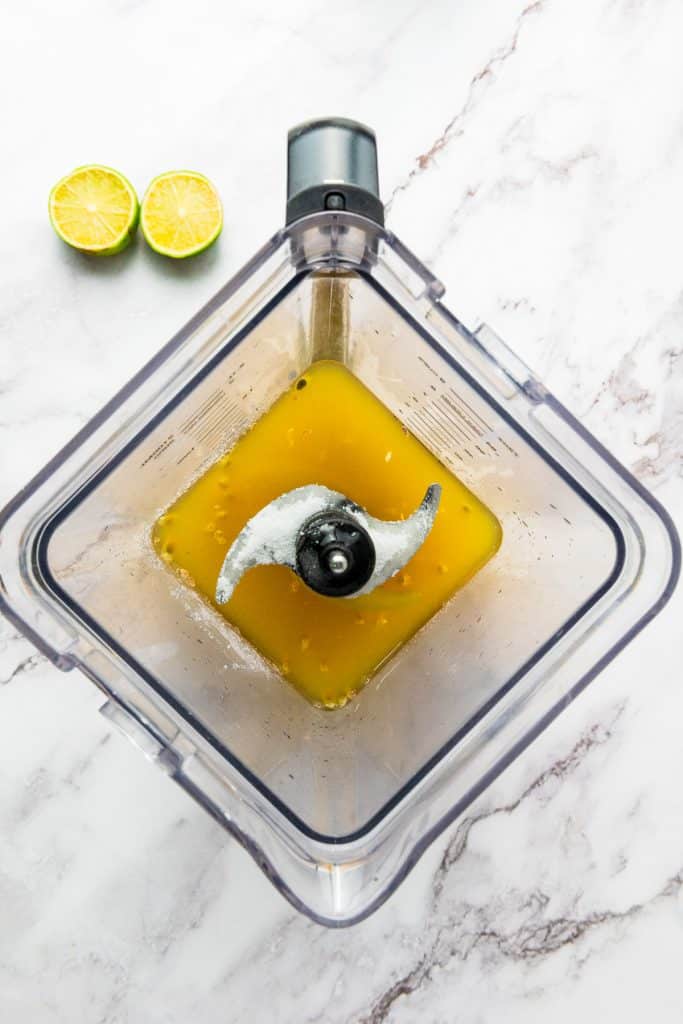 Overhead view of a blender with orange passion fruit juice and ice, next to two squeezed lime halves on a marble surface.