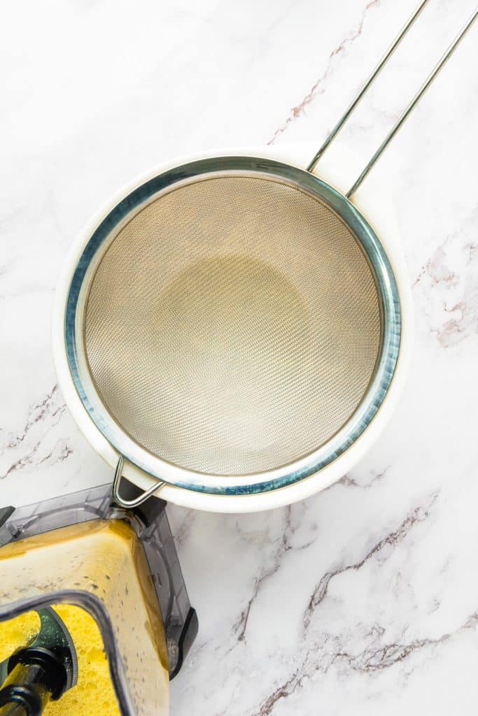 A blender with yellow passion fruit juice sits near a mesh strainer over a white bowl on a marble counter.