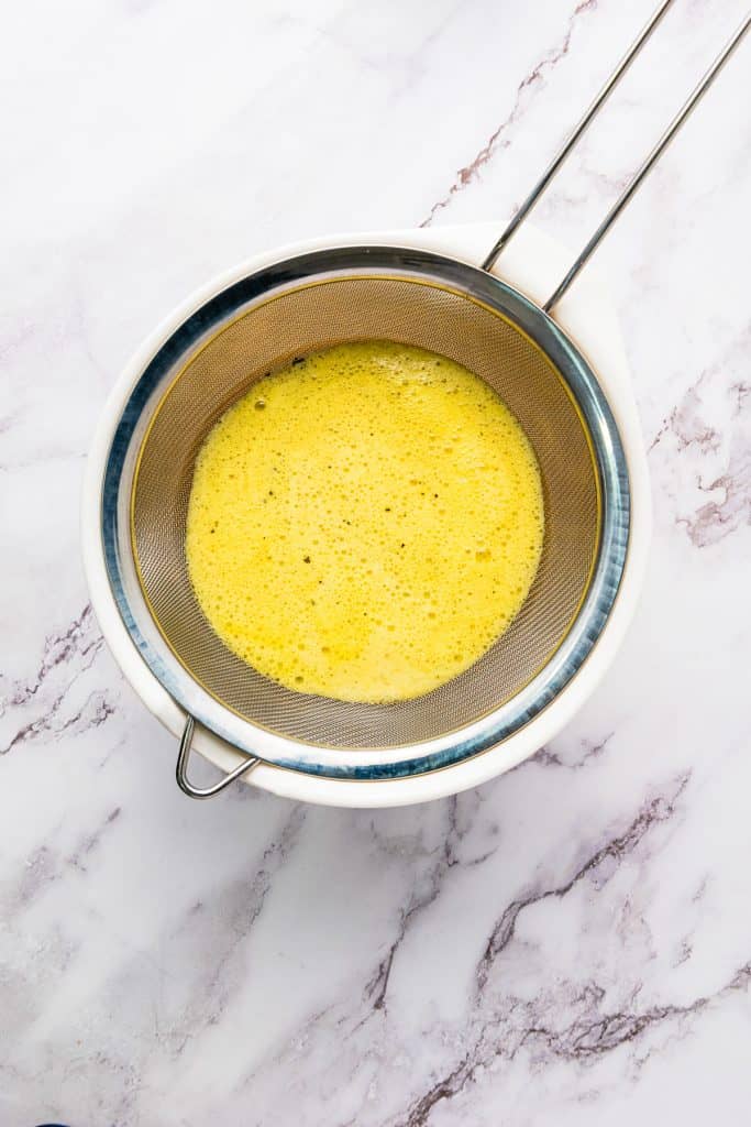 Metal strainer over a white bowl with vibrant passion fruit juice being strained, on a marble surface.