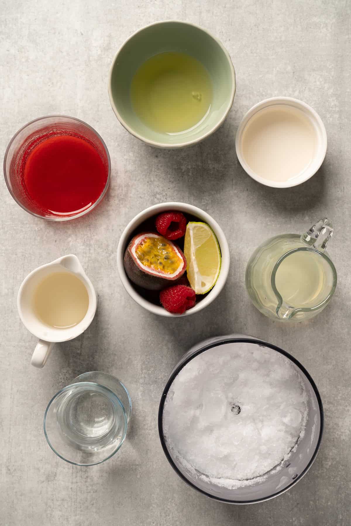 Ingredients for a raspberry passionfruit tequila sour mocktail, including juice, lime, berries, syrup, and crushed ice, arranged in bowls and cups.