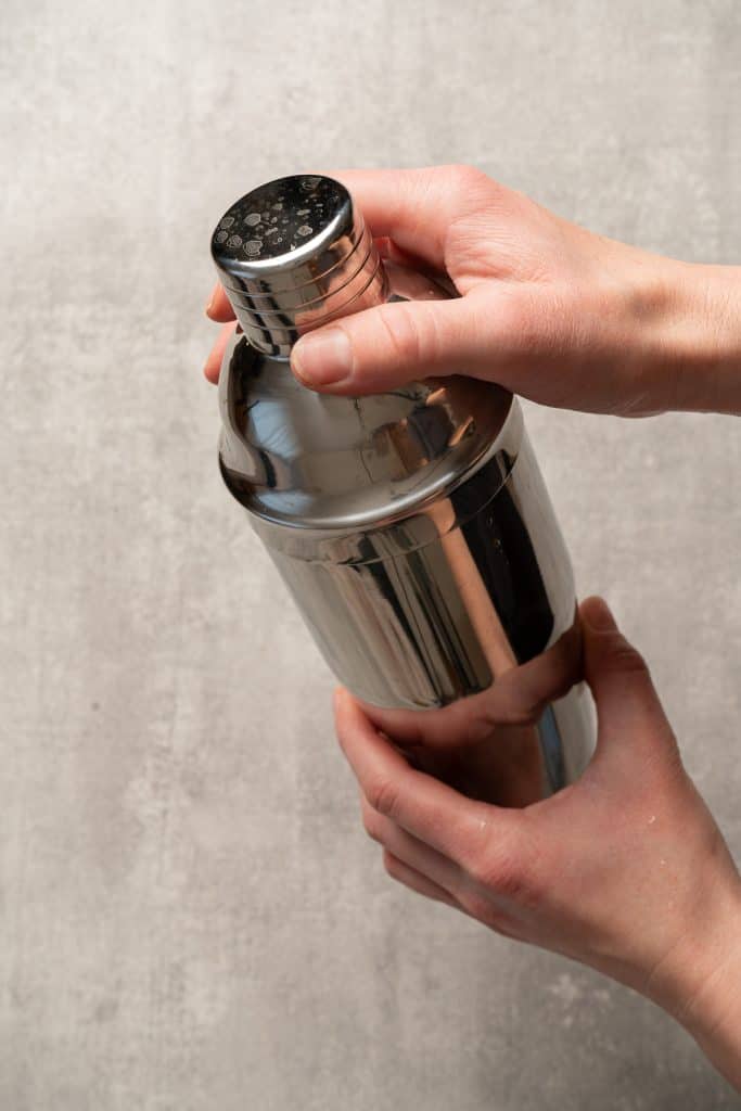 Two hands holding a stainless steel cocktail shaker against a light grey background, ready to mix a refreshing raspberry passionfruit tequila sour mocktail.