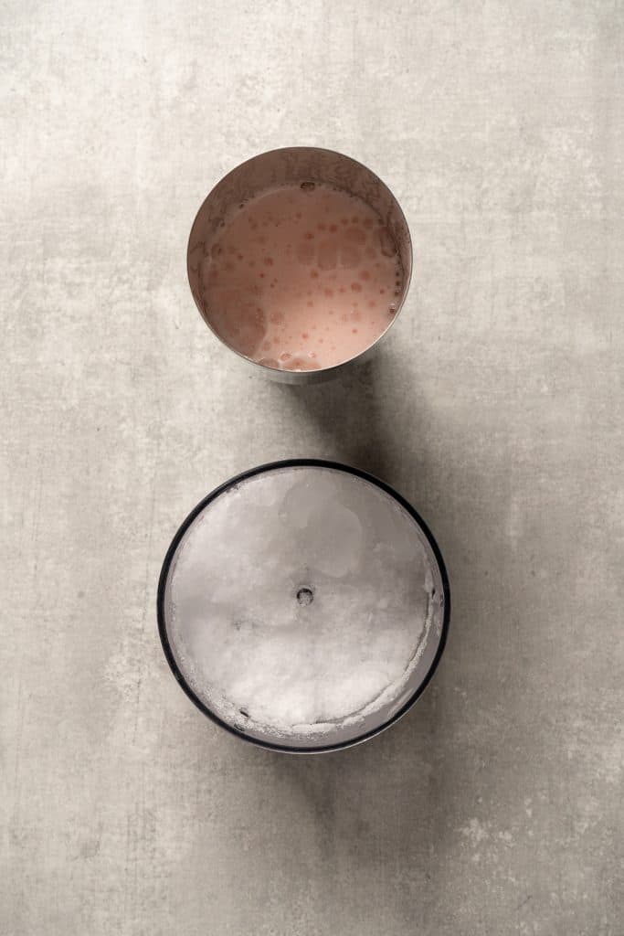 A metal cup with pink frothy raspberry passionfruit tequila sour mocktail beside a cup filled with crushed ice, on a gray surface.