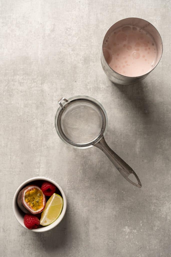 A cocktail shaker, a strainer, and a bowl with passion fruit, raspberries, and lime on a gray surfaceโperfect ingredients for crafting a refreshing raspberry passionfruit tequila sour mocktail.