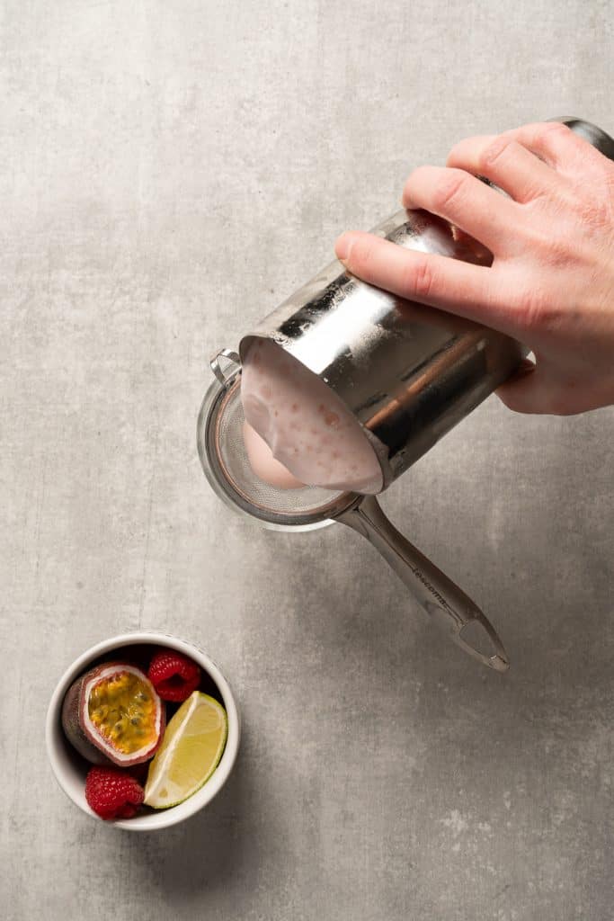 A hand pours a raspberry passionfruit tequila sour mocktail from a shaker next to a bowl with passionfruit, raspberries, and a lime wedge.