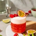 A vibrant raspberry passionfruit tequila sour mocktail garnished with raspberries and half a passion fruit sits on a board with limes nearby.