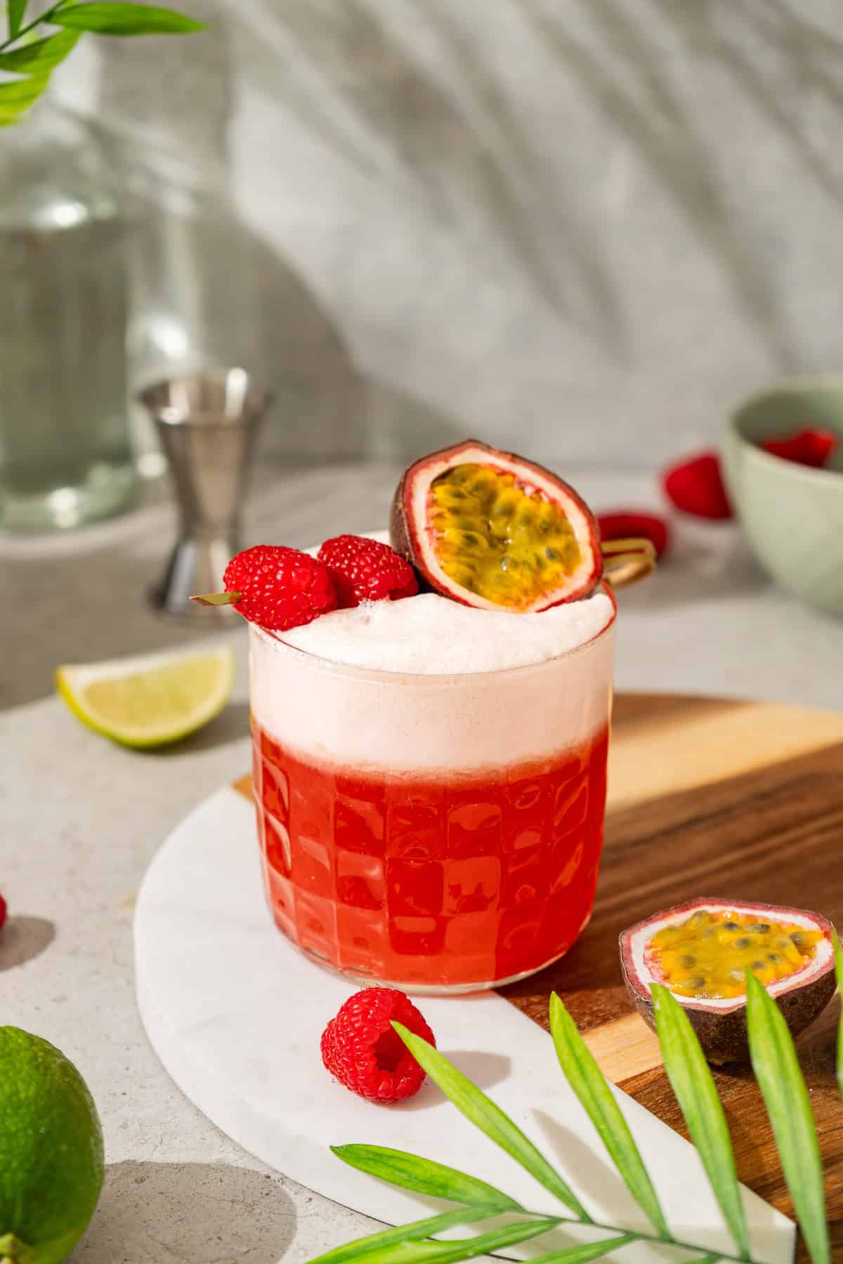 A vibrant raspberry passionfruit tequila sour mocktail garnished with raspberries and half a passion fruit sits on a board with limes nearby.