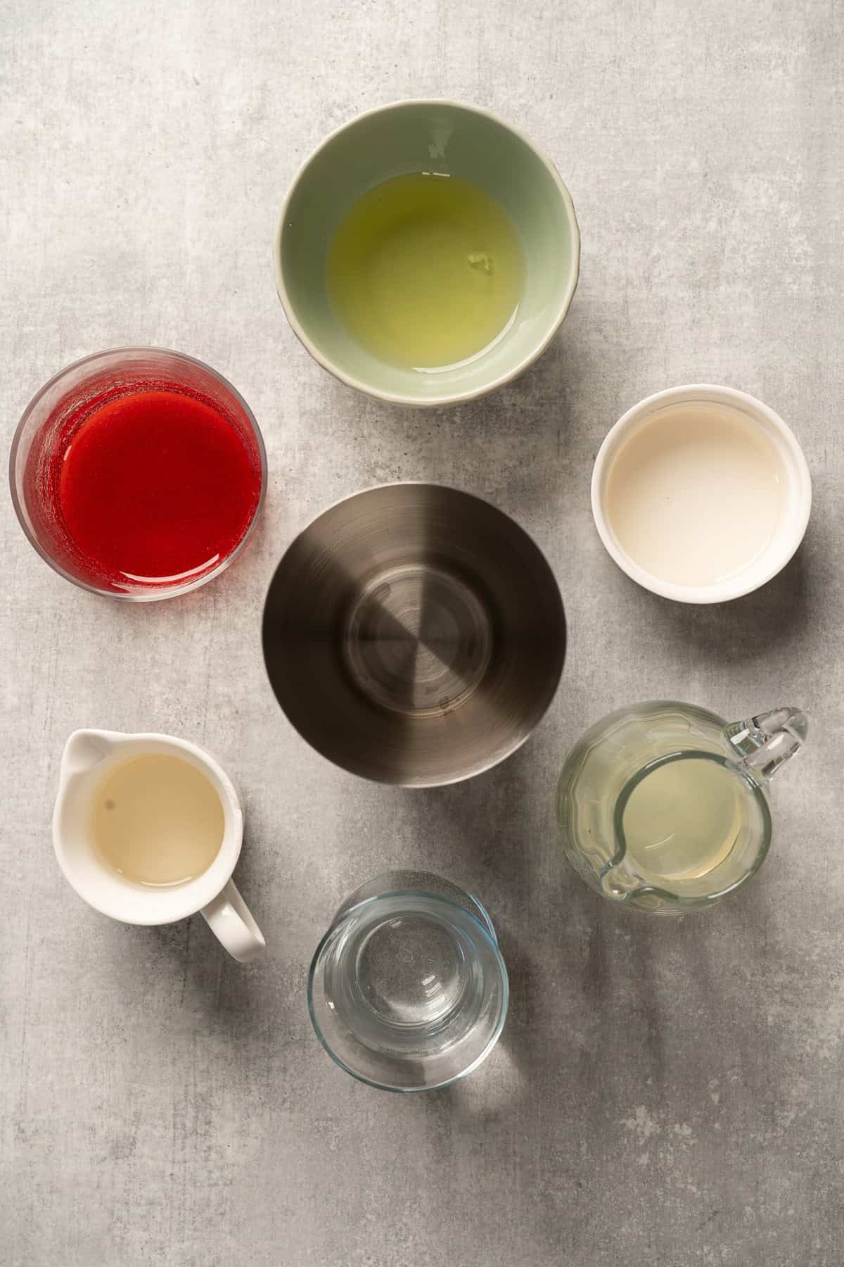 Seven bowls and pitchers containing various liquidsโincluding a vibrant raspberry passionfruit tequila sour mocktailโarranged in a circle on a gray surface, viewed from above.