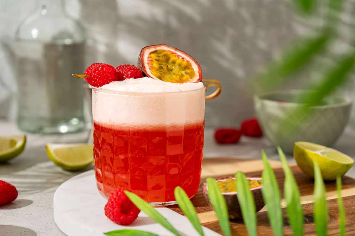 A frothy red raspberry passionfruit tequila sour mocktail garnished with raspberries and a passion fruit half, with limes and leaves nearby.