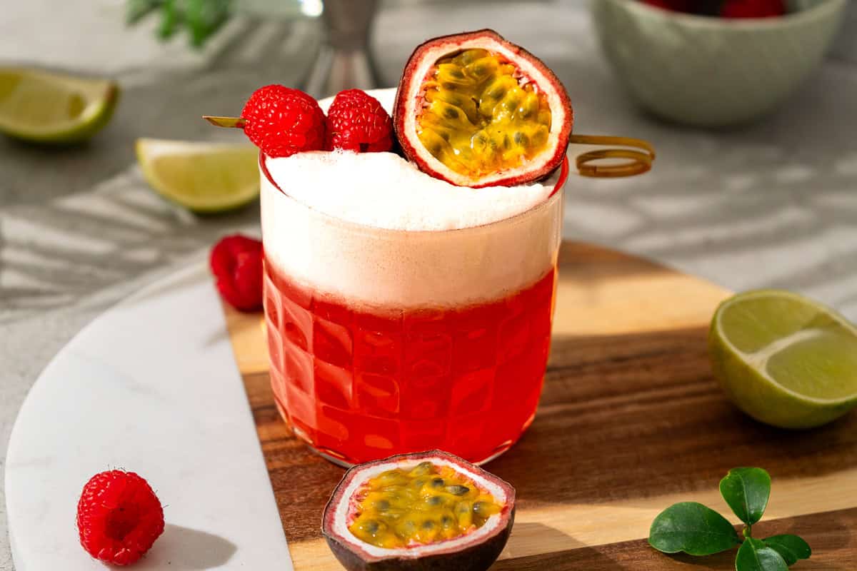A raspberry passionfruit tequila sour mocktail, this red cocktail with foam is garnished with raspberries and half a passion fruit, served on a wooden board with lime slices.