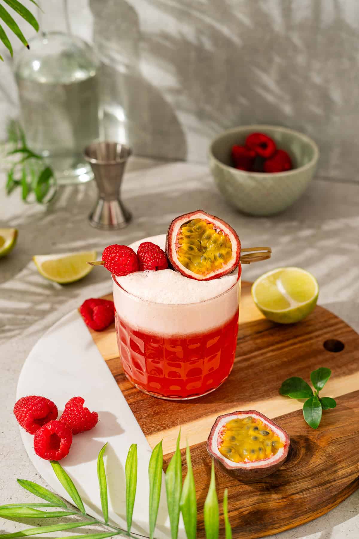 A vibrant pink raspberry passionfruit tequila sour mocktail sits on a wooden board, garnished with raspberries, passion fruit, limes, and berries.