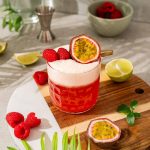 A vibrant raspberry passionfruit tequila sour mocktail sits on a board, garnished with raspberries and passion fruit, surrounded by fresh fruit.