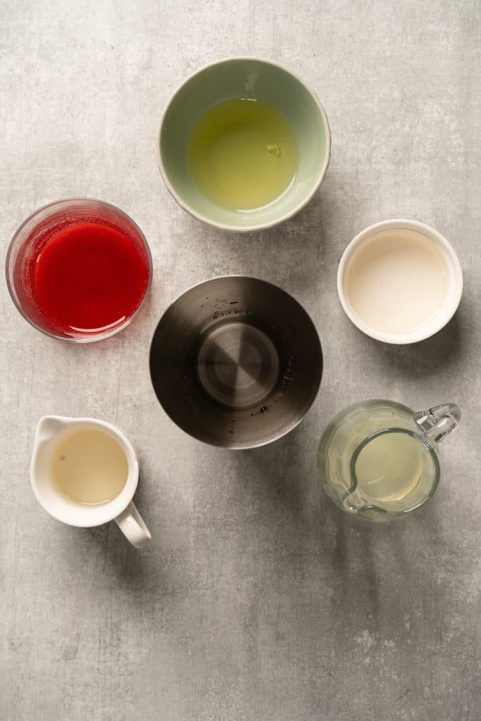 Six bowls and pitchers containing various liquids, such as a vibrant raspberry passionfruit tequila sour mocktail, arranged on a light gray surface, viewed from above.