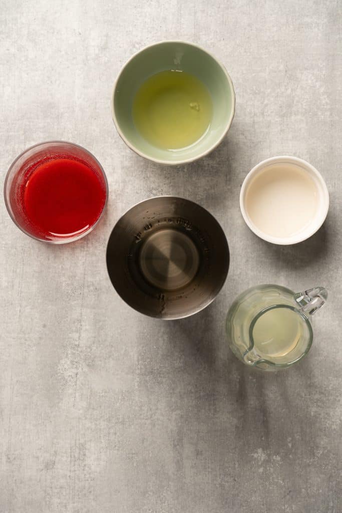 Five bowls with different liquids, including one resembling a raspberry passionfruit tequila sour mocktail, are arranged on a light gray surface and viewed from above.