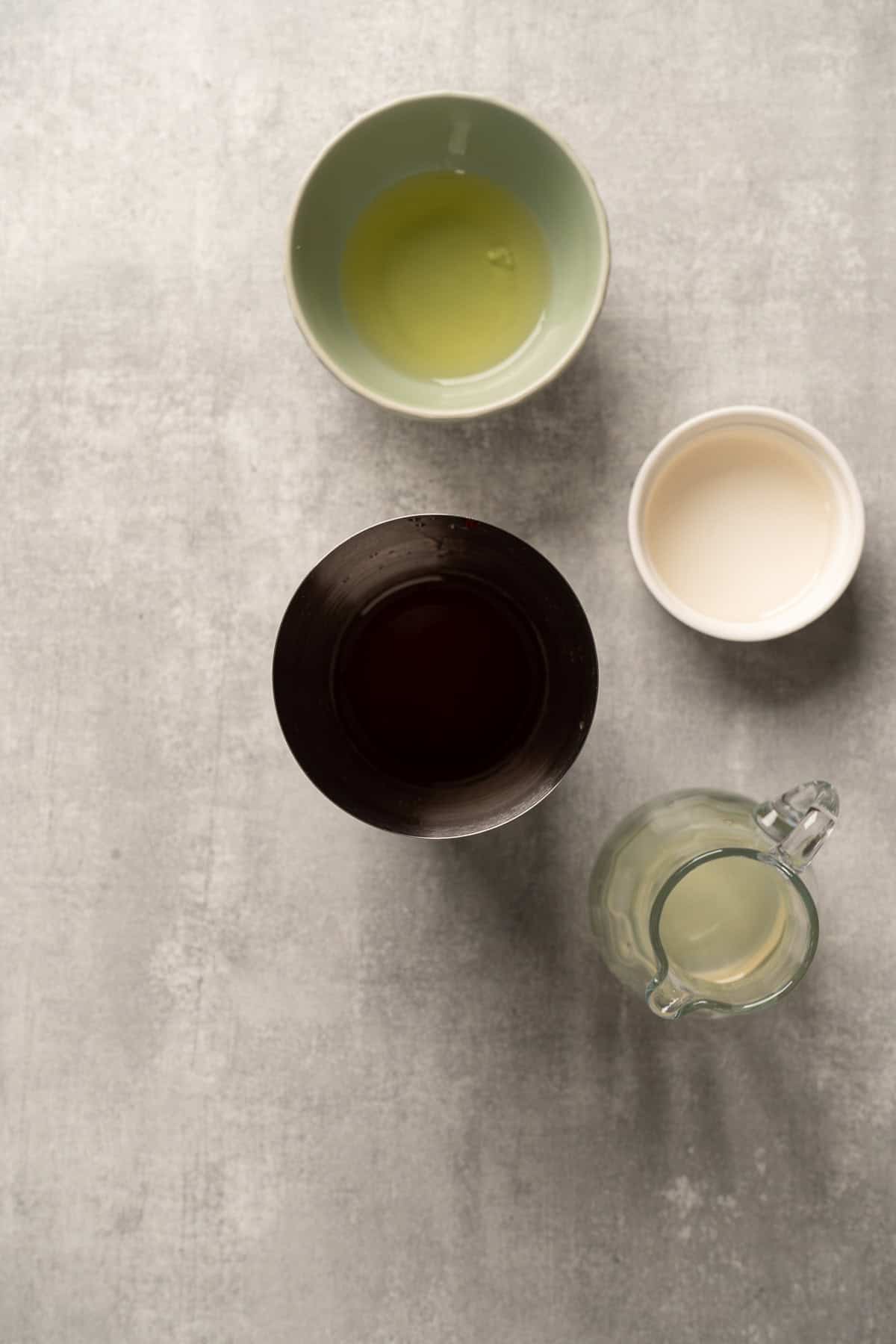 Four bowls and a jug with different clear liquids, perfect for crafting a raspberry passionfruit tequila sour mocktail, are arranged on a light gray surface and viewed from above.