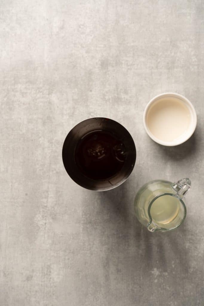 Three containers viewed from above: a dark glass, a small white bowl, and a clear glass jug filled with raspberry passionfruit tequila sour mocktail.
