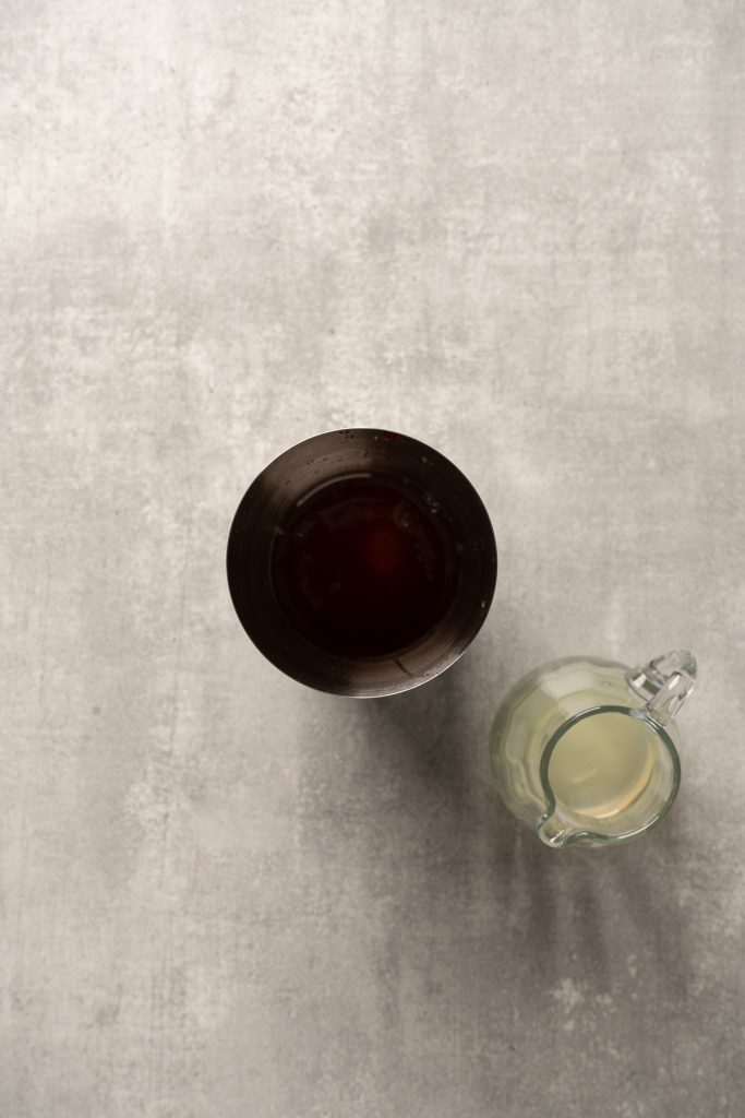 A raspberry passionfruit tequila sour mocktail in a dark glass sits next to a small glass pitcher of milk on a light gray surface.