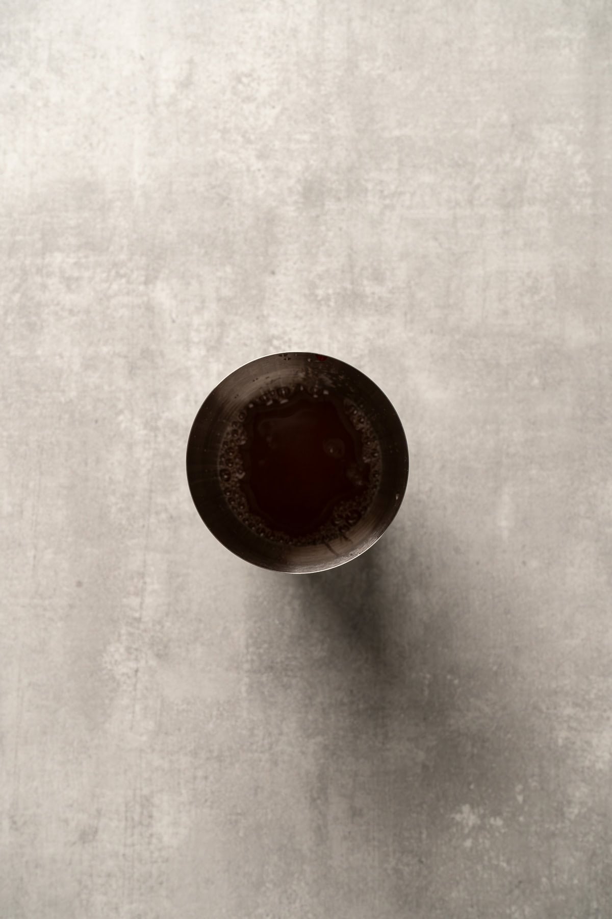 Aerial view of a glass filled with dark liquid on a light gray textured surface, showcasing a refreshing raspberry passionfruit tequila sour mocktail.