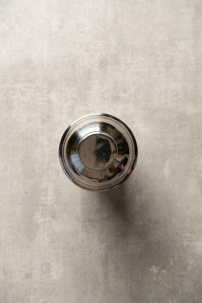 A stainless steel cocktail shaker viewed from above on a light gray textured surface, ready to mix a vibrant raspberry passionfruit tequila sour mocktail.