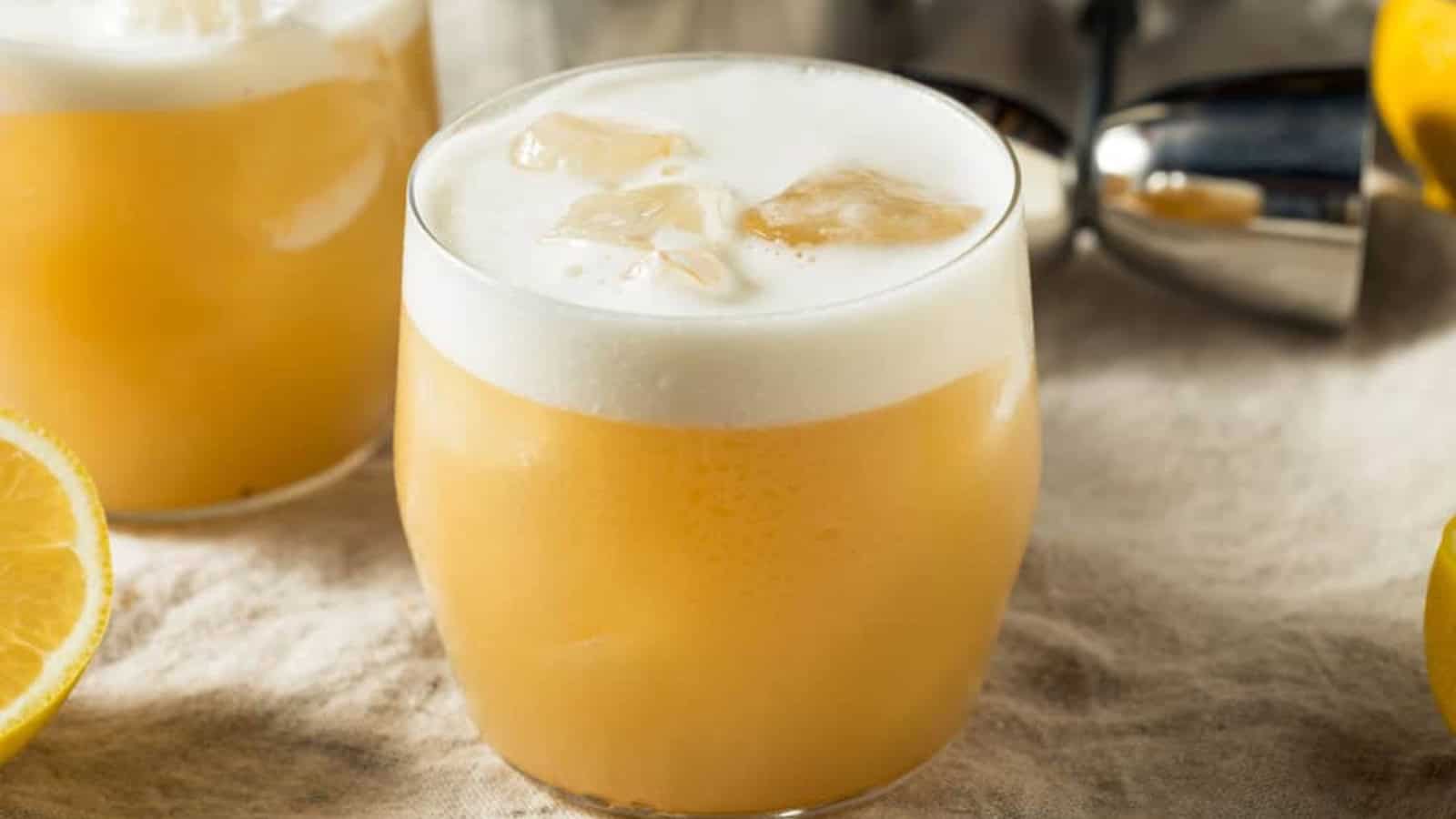 A glass of whiskey sour with foam on top, ice cubes, and a lemon slice nearby on a light cloth surface.