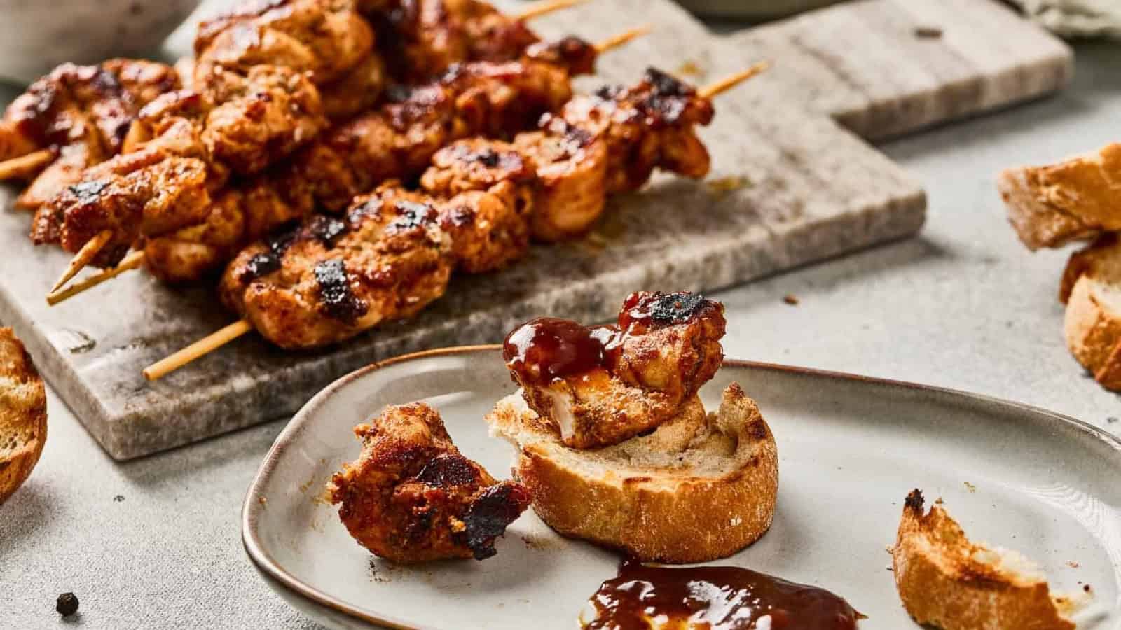 Grilled chicken skewers with spices on a cutting board, with sauce and bread nearby.