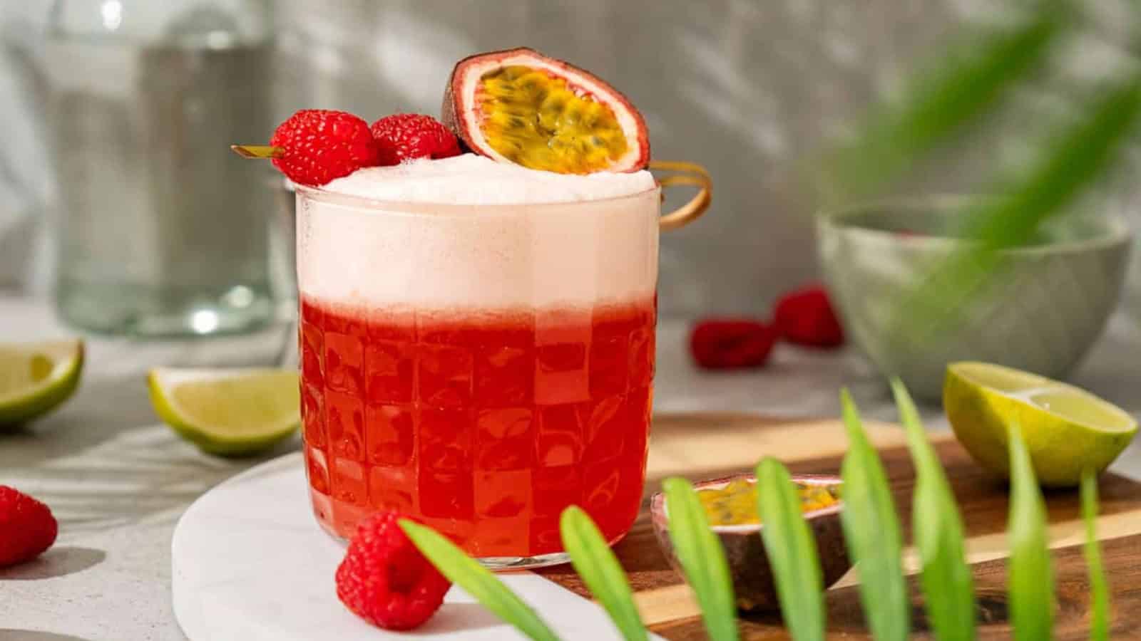 A red cocktail with foam, garnished with raspberries and half a passion fruit, on a wooden tray with lime slices.