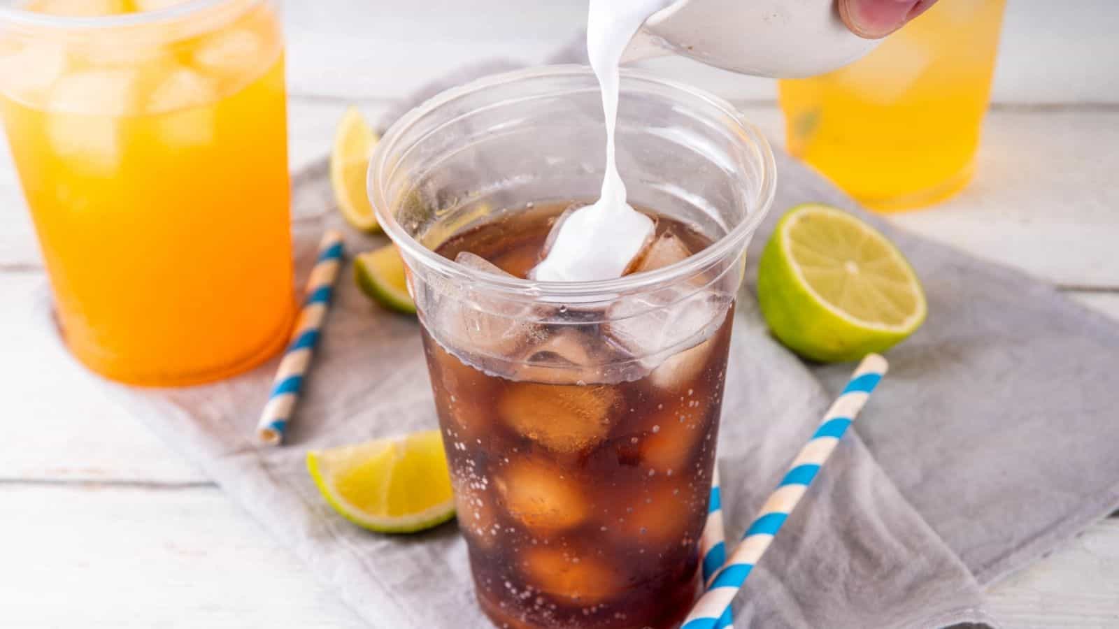 Cream being poured into an iced cola drink in a clear cup, with lime wedges and striped straws nearby.