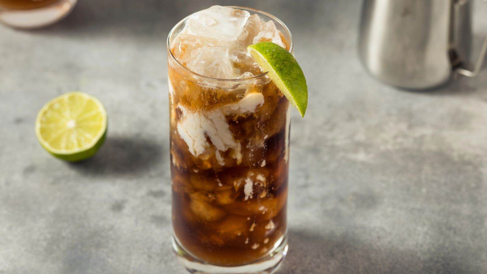A glass of iced dark drink with a lime wedge, next to a sliced lime on a gray surface.