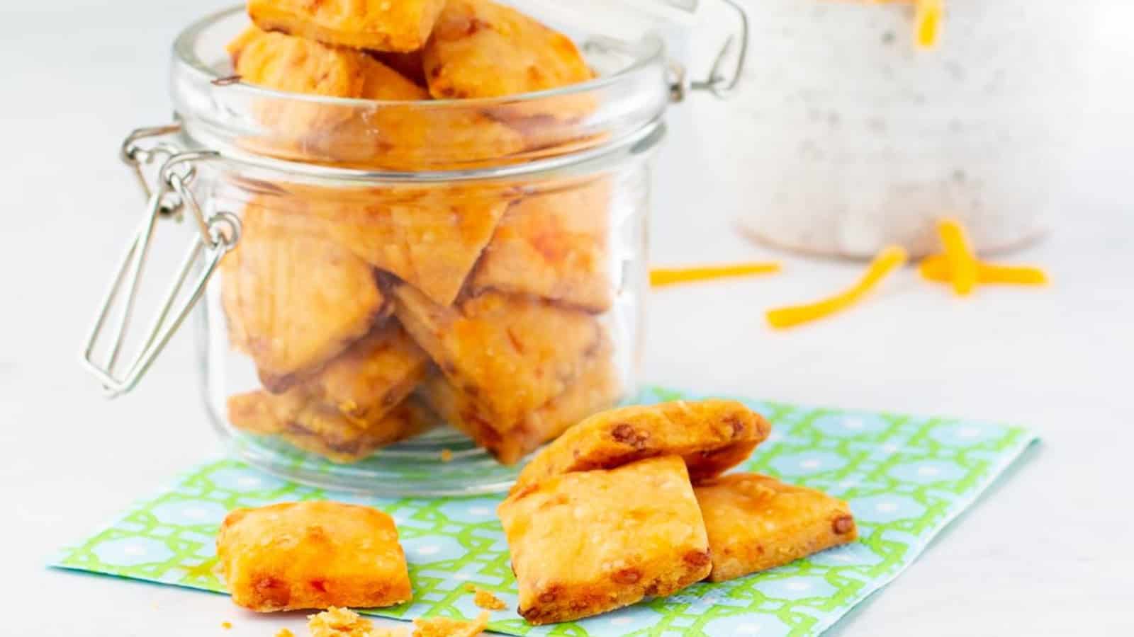 Square cheddar crackers spill from a glass jar onto a napkin, with cheese shreds in the background.