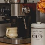 Espresso machine with mug, coffee canister, and a vase of orange flowers on a wooden countertop-perfect inspiration for your home café or stylish coffee bar ideas.