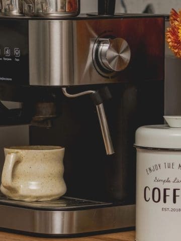 Espresso machine with mug, coffee canister, and a vase of orange flowers on a wooden countertopโperfect inspiration for your home cafรฉ or stylish coffee bar ideas.