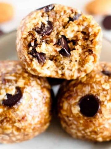Three almond chocolate chip energy balls in a bowl, surrounded by almonds and chocolate chips on a white surface.