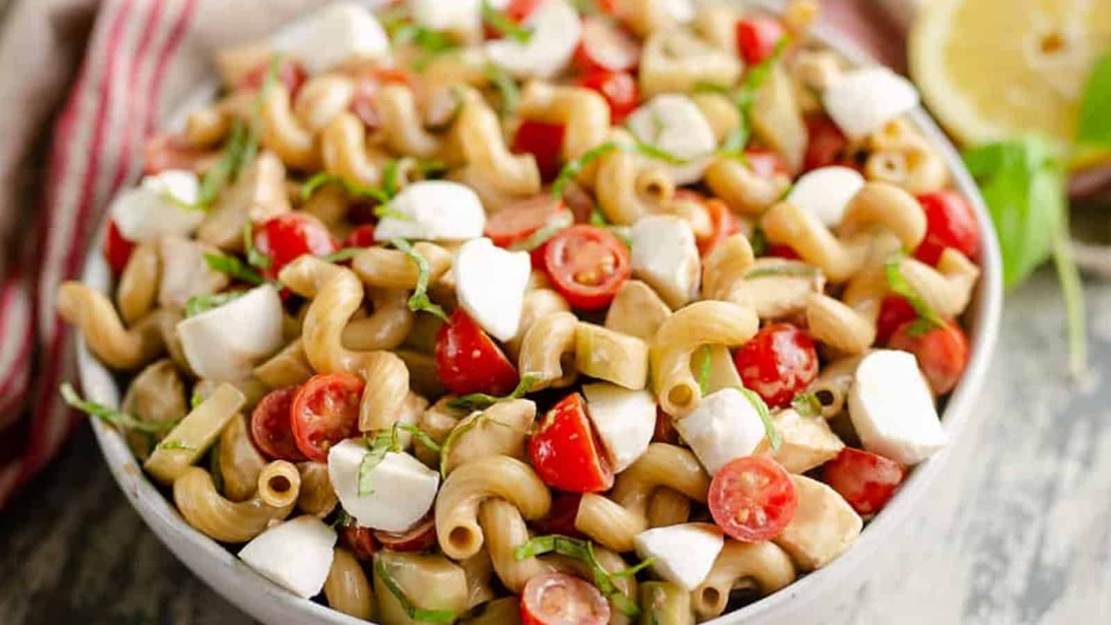 Bowl of pasta salad with cherry tomatoes, mozzarella, and fresh basil, garnished with lemon in the background.