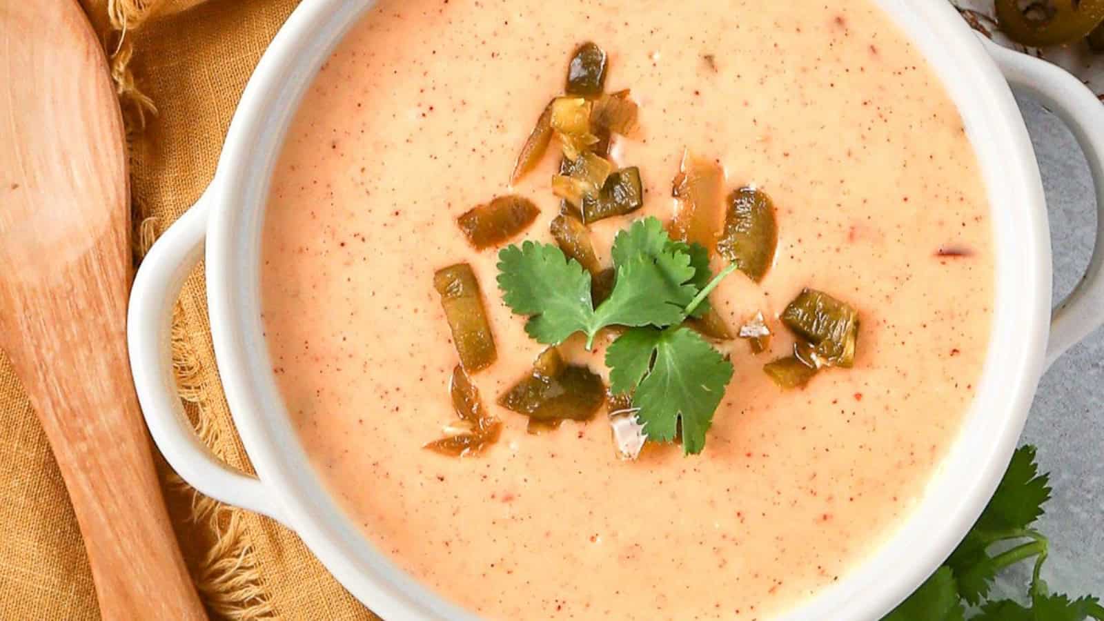 Creamy queso dip topped with chopped green peppers and fresh cilantro in a white bowl, beside a wooden spoon.