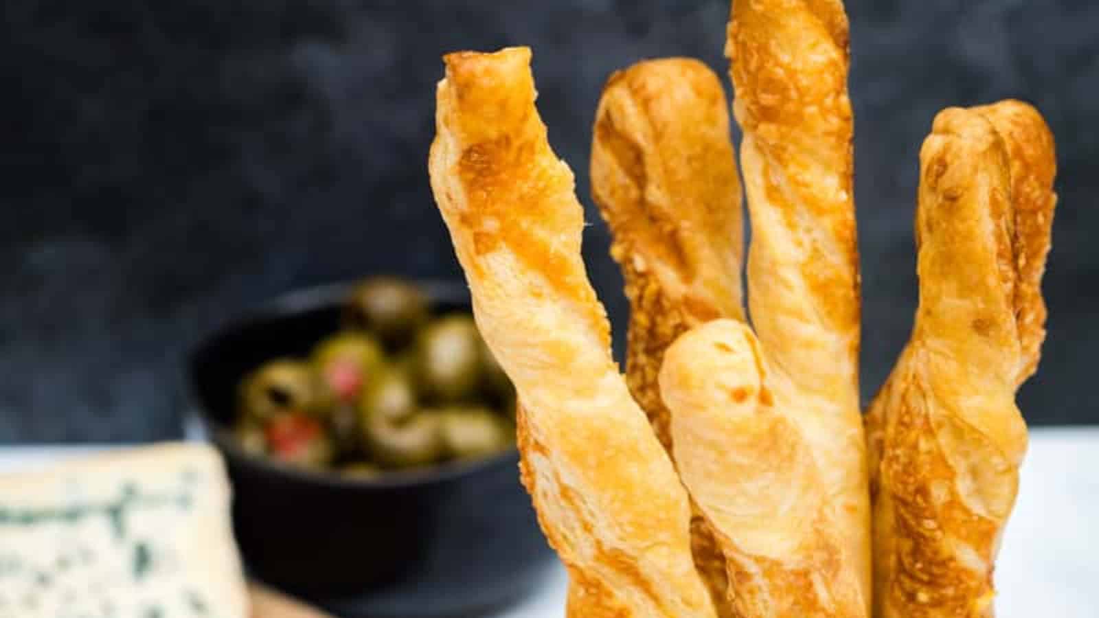 Four cheese straws stand upright with a bowl of green olives blurred in the background.