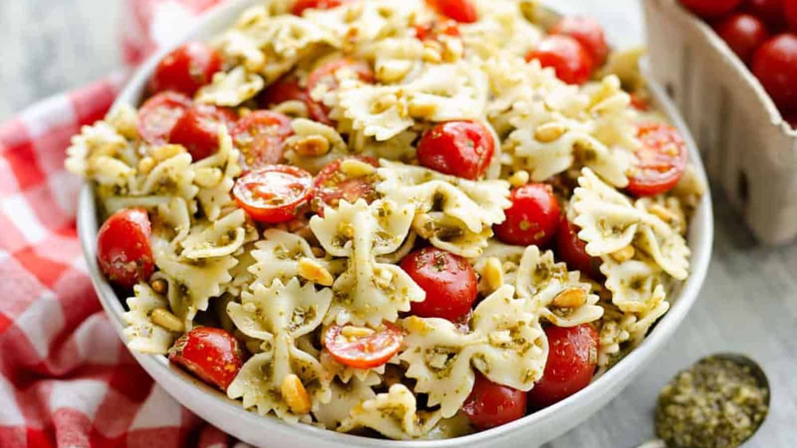 Bowl of bow tie pasta with pesto, cherry tomatoes, and pine nuts on a red checkered cloth.