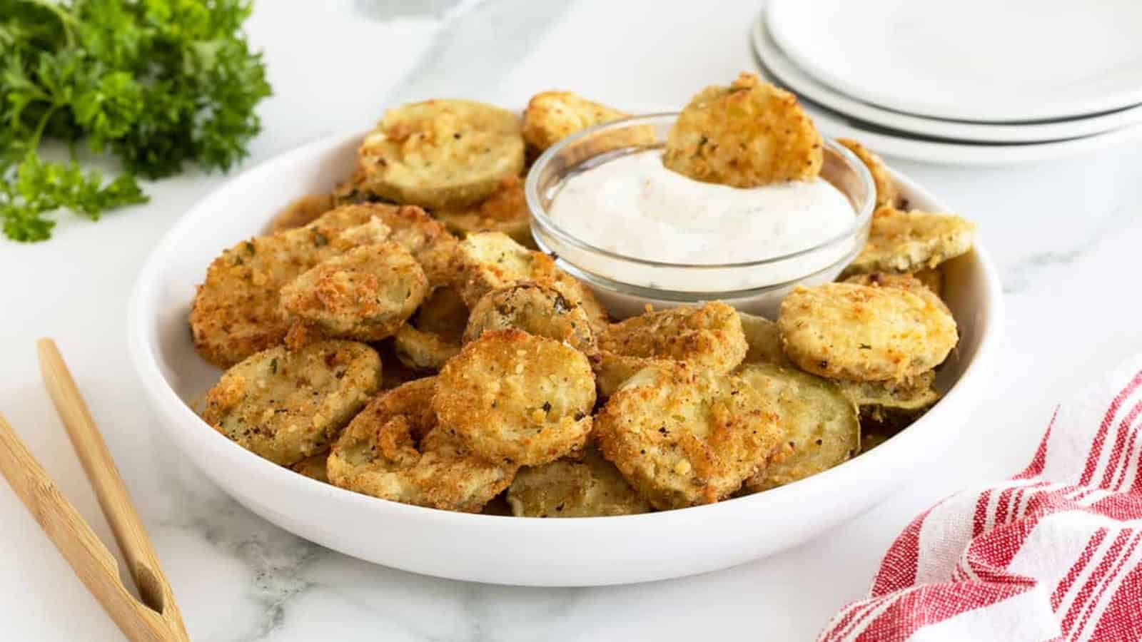 A plate of fried pickles with a bowl of dipping sauce sits on a table next to a red-striped towel.