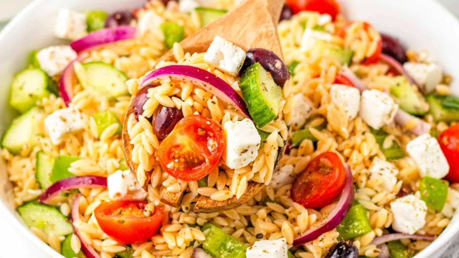 A wooden spoon lifts a serving of orzo pasta salad with cherry tomatoes, cucumber, red onion, feta cheese, and olives from a white bowl.
