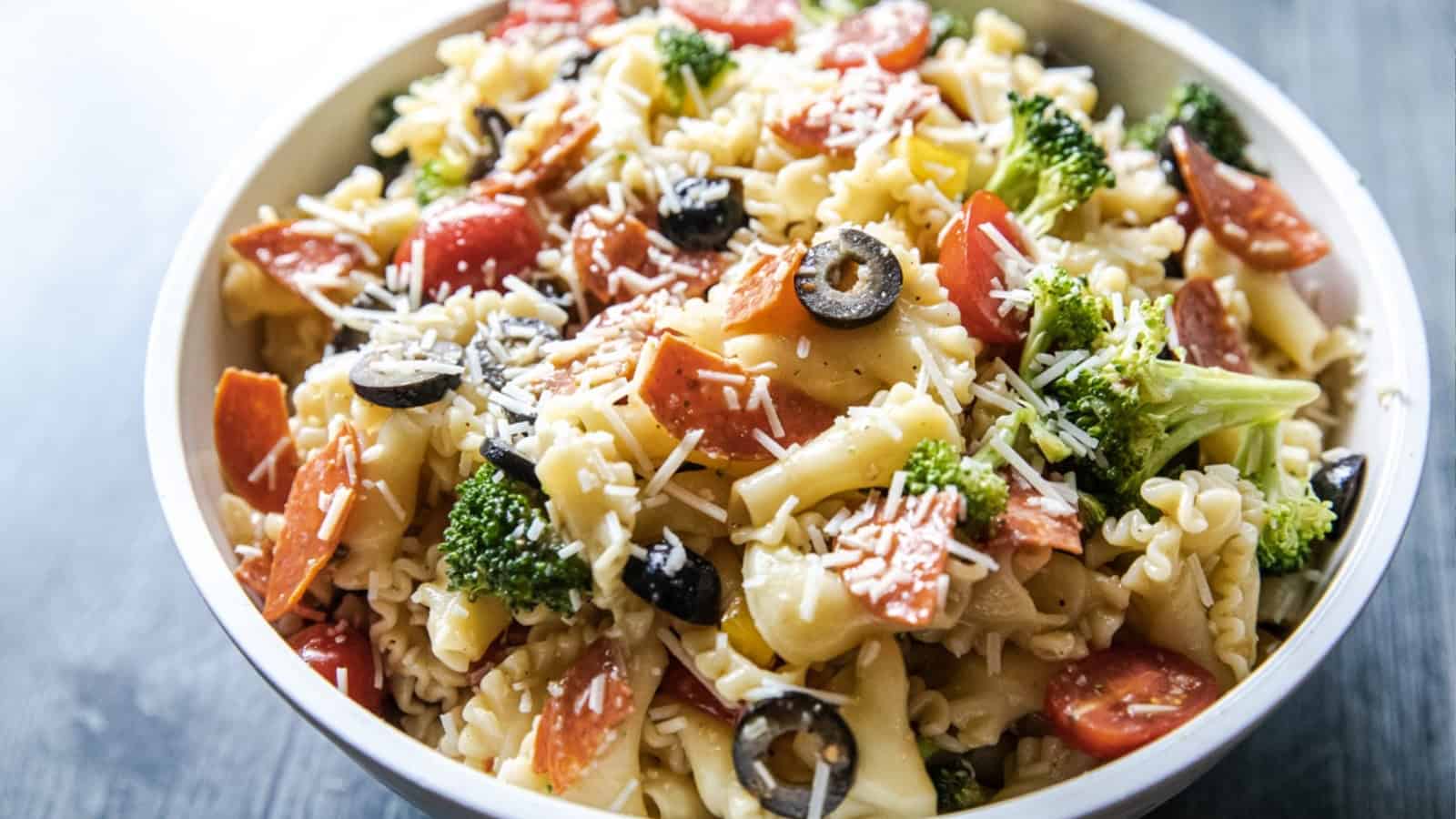 A bowl of pasta salad with broccoli, tomatoes, olives, pepperoni, and shredded cheese.