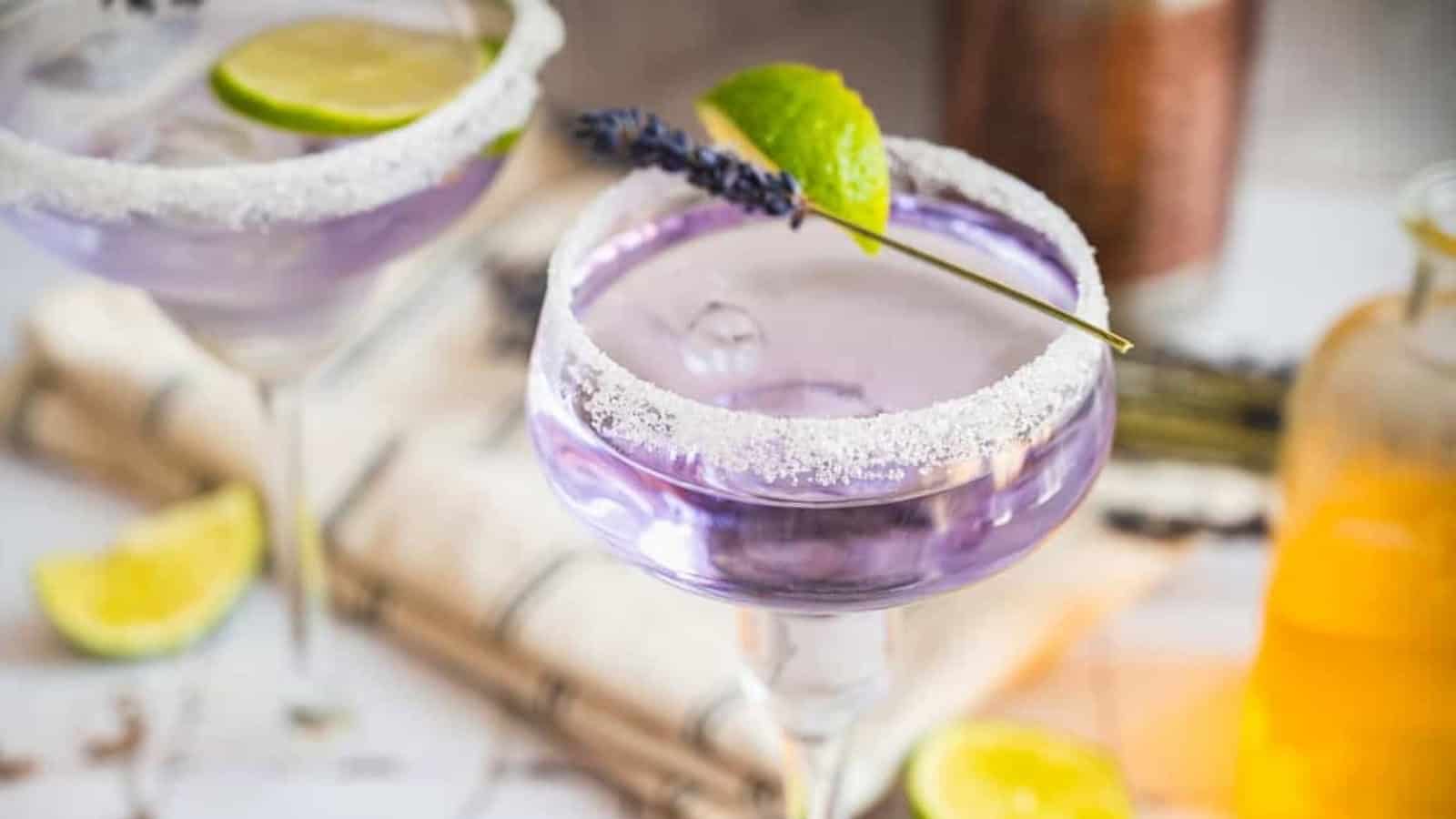 A purple cocktail in a sugar-rimmed glass, garnished with a lime wedge and a sprig of lavender.