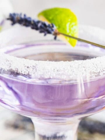 Purple cocktail in a sugar-rimmed glass, garnished with a lime wedge and lavender sprig.