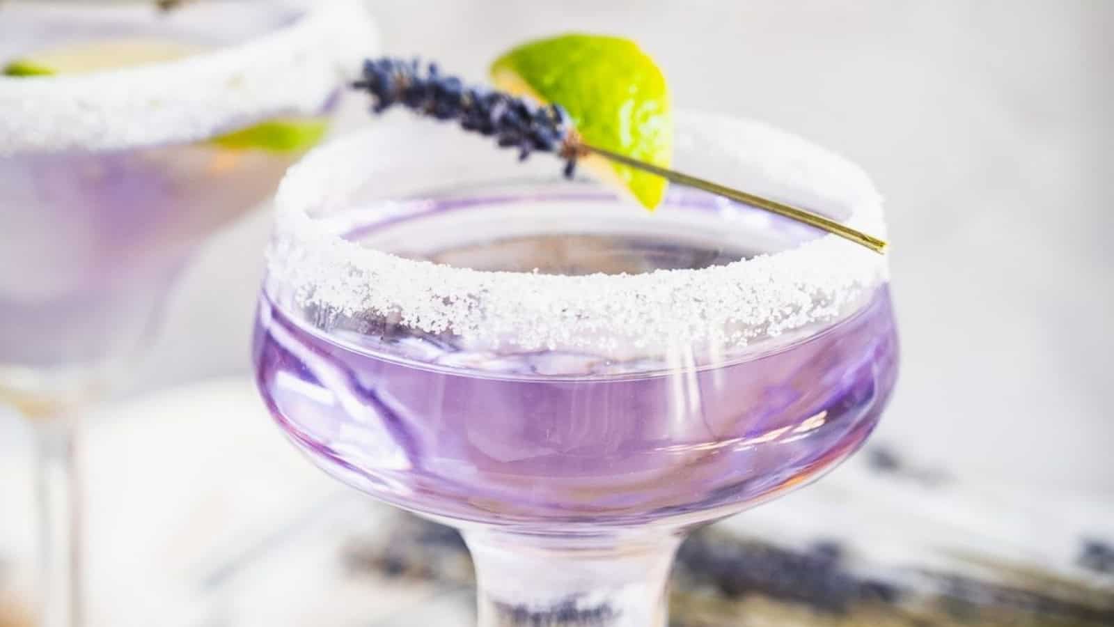 A purple cocktail in a sugar-rimmed glass, garnished with a lime wedge and a sprig of lavender.