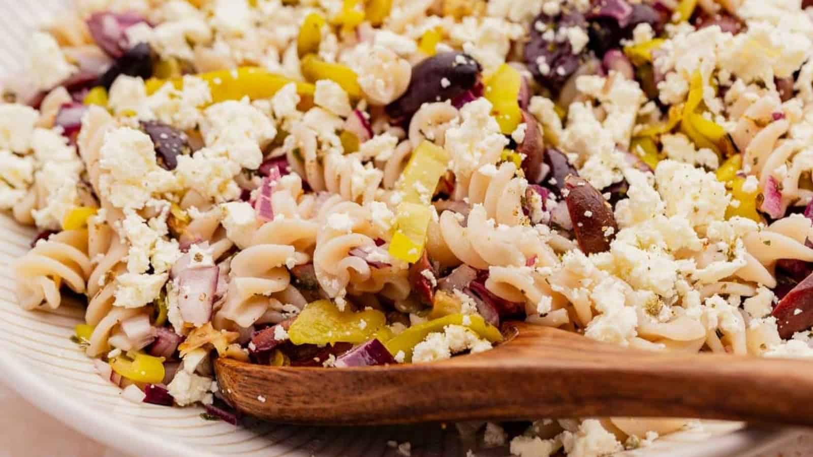 Rotini pasta salad with feta cheese, olives, peppers, and onions, served with a wooden spoon on a plate.