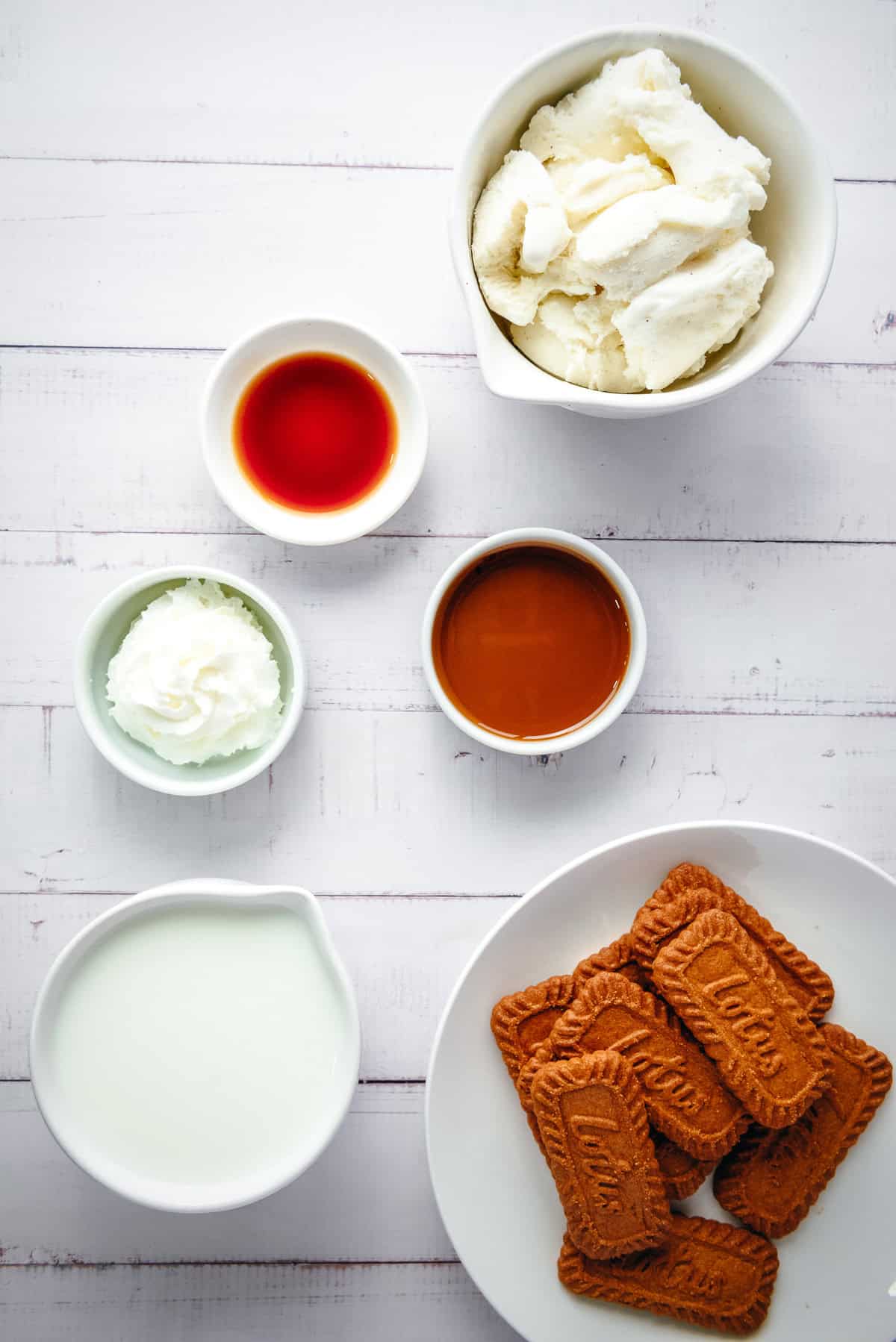 Bowls of cream, milk, biscoff cookies, caramel, vanilla, and whipped cream on a white wooden tableโperfect for creating a delicious Biscoff dessert or a creamy Biscoff milkshake.